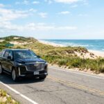 Luxury black Cadillac Escalade driving along scenic coastal road towards the Hamptons with ocean dunes and bright summer sun